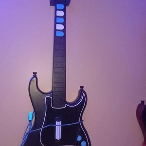 Op de afbeelding: Zwarte gitaarvormige gamecontroller met witte randen en blauwe accenten. De controller heeft een hals met blauwe en witte knoppen en de merknaam "Kramer" op de kop. Een blauwe kabel is aangesloten op de body.