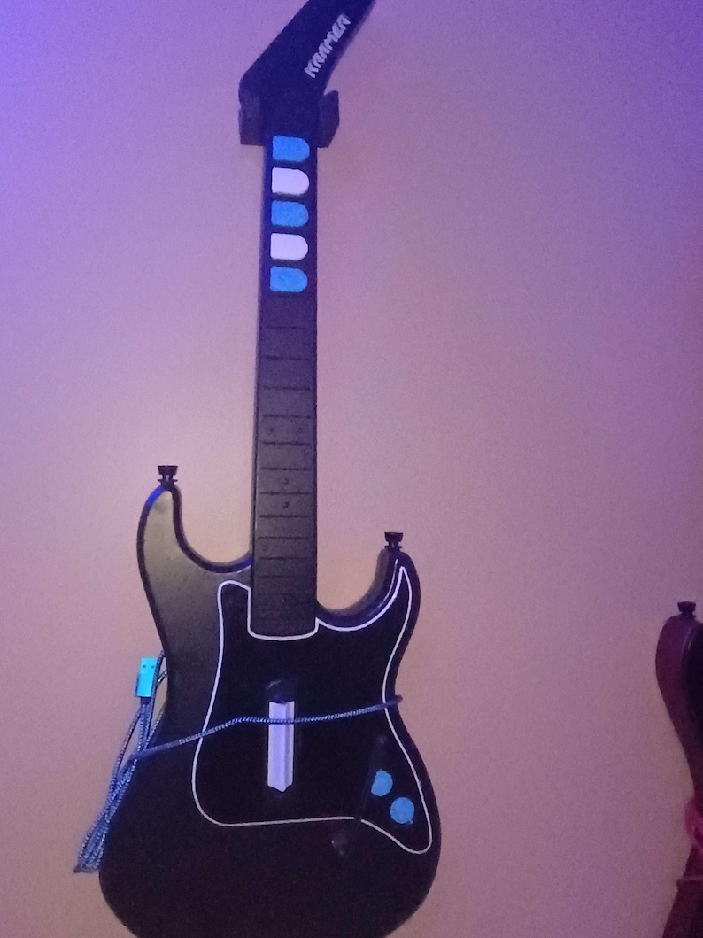 Op de afbeelding: Zwarte gitaarvormige gamecontroller met witte randen en blauwe accenten. De controller heeft een hals met blauwe en witte knoppen en de merknaam "Kramer" op de kop. Een blauwe kabel is aangesloten op de body.