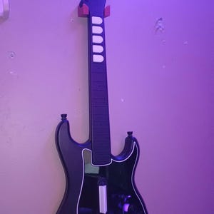 Op de afbeelding: Zwarte Kramer gitaarcontroller voor een videogame, aan de muur bevestigd. De gitaar heeft een witte omtrek en witte fretknoppen. De kop is zwart met het Kramer-logo.