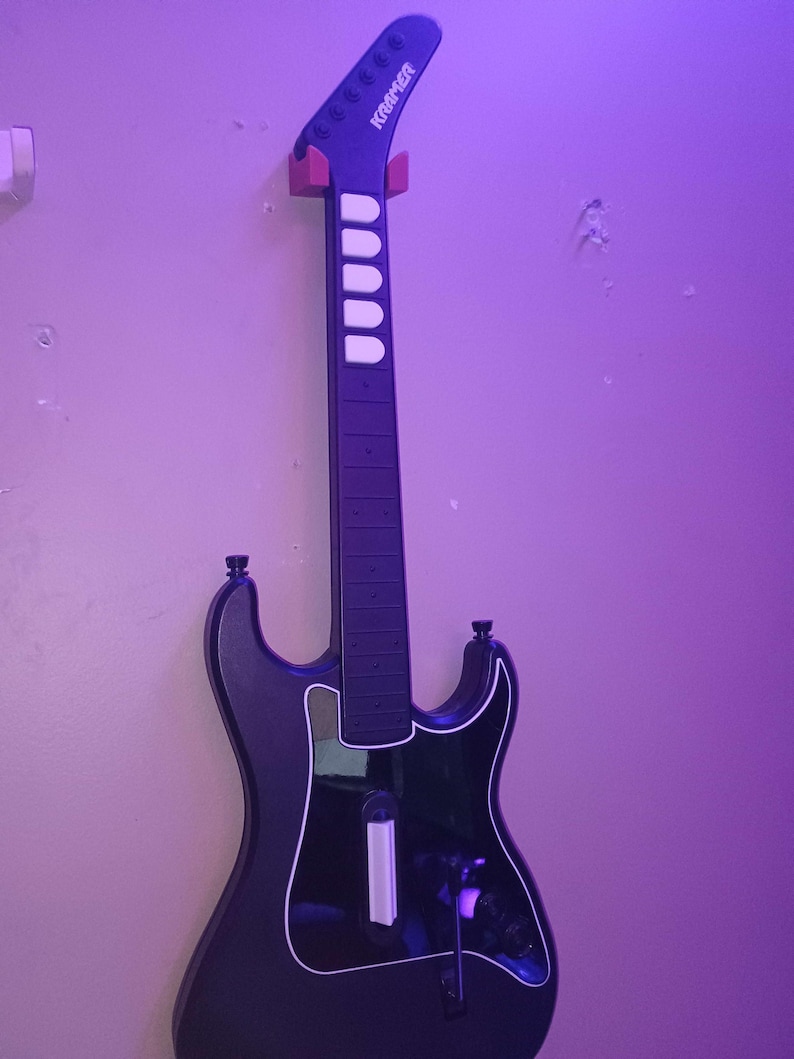 Op de afbeelding: Zwarte Kramer gitaarcontroller voor een videogame, aan de muur bevestigd. De gitaar heeft een witte omtrek en witte fretknoppen. De kop is zwart met het Kramer-logo.
