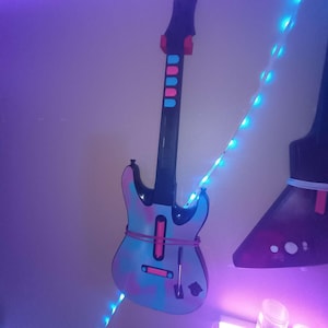 May include: A decorative guitar-shaped wall hanging with a blue, pink, and purple design. The guitar has a black neck and headstock with red and blue accents. A string of blue lights is wrapped around the guitar.