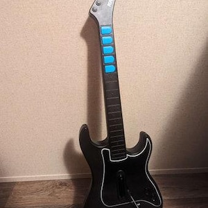 Op de afbeelding: Zwarte gitaarvormige gamecontroller met een witte omtrek en blauwe knoppen. De kop is grijs met de merknaam "Kramer" erop gedrukt. De controller leunt tegen een muur en een houten vloer.