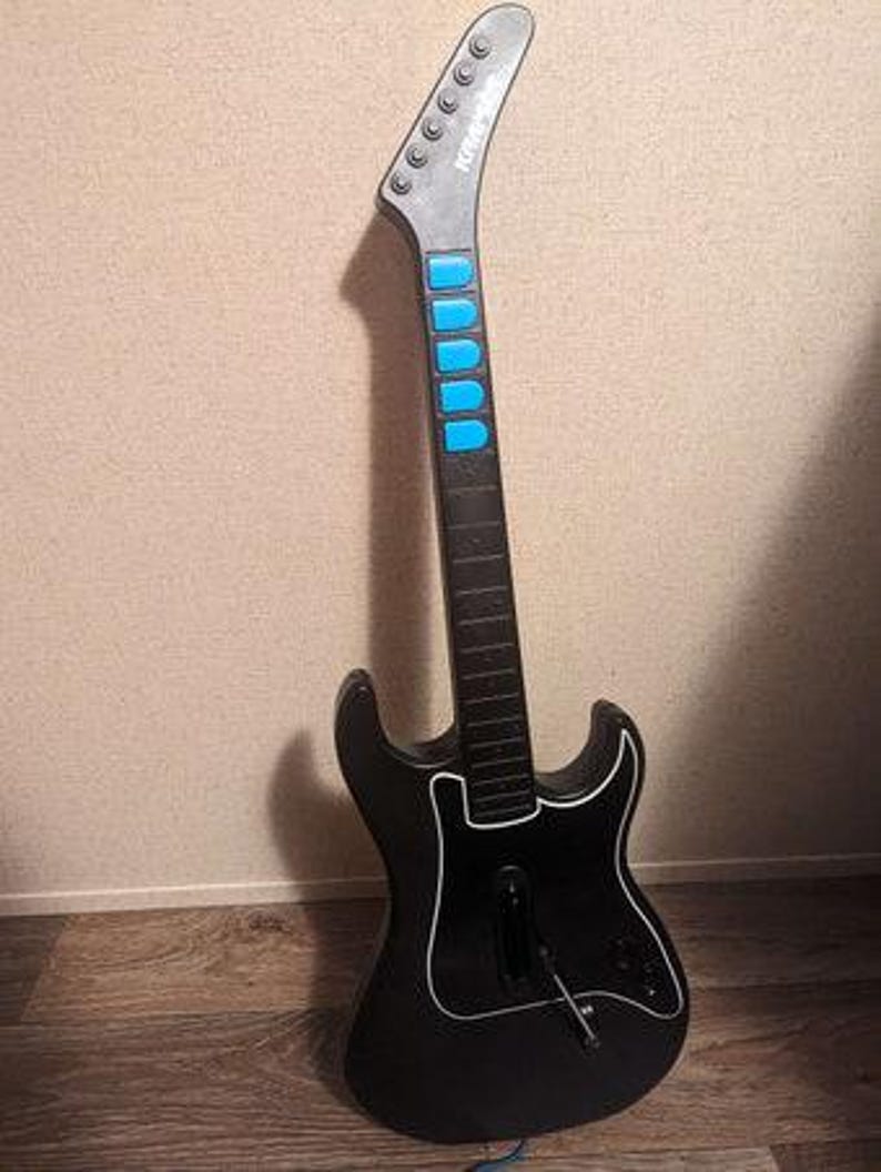 Op de afbeelding: Zwarte gitaarvormige gamecontroller met een witte omtrek en blauwe knoppen. De kop is grijs met de merknaam "Kramer" erop gedrukt. De controller leunt tegen een muur en een houten vloer.