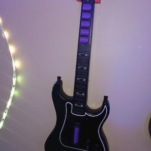 Op de afbeelding: Zwarte speelgoedgitaar met een witte omtrek en paarse knoppen. De gitaar heeft een zwarte hals met zilveren frets en een kop met het woord "Kramer" in het wit. Een rode houder ondersteunt de gitaar aan de muur.