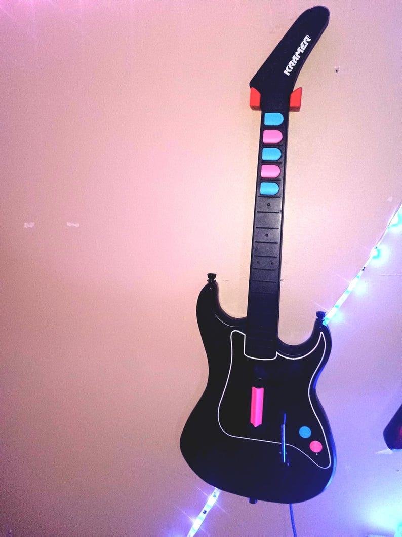 Op de afbeelding: Zwarte Kramer gitaarvormige gamecontroller met een witte omtrek. De hals heeft roze en blauwe knoppen. De kop is zwart met het woord "Kramer" in het wit. De controller is aan een muur bevestigd.
