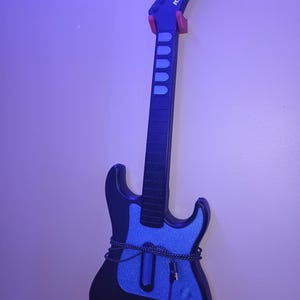 Op de afbeelding: Een zwarte en blauwe Kramer gitaar met een blauwe glitterafwerking op de body. De gitaar hangt aan een muur met een rode gitaarhouder. De kop heeft het Kramer logo.
