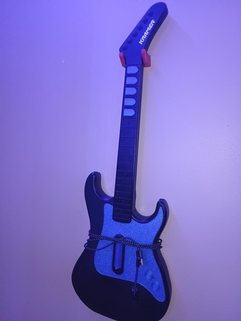 Op de afbeelding: Een zwarte en blauwe Kramer gitaar met een blauwe glitterafwerking op de body. De gitaar hangt aan een muur met een rode gitaarhouder. De kop heeft het Kramer logo.