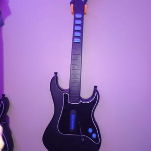 Op de afbeelding: Zwarte Kramer gitaarvormige gamecontroller die aan een muur hangt. De controller heeft een witte omtrek, blauwe knoppen en een blauw licht. De kop heeft het Kramer-logo en wordt vastgehouden door oranje steunen.