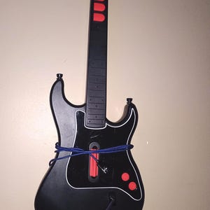 Op de afbeelding: Een zwart-rode gitaarvormige videogamecontroller met witte rand. De controller heeft rode knoppen en een blauw koord eromheen gewikkeld. De bovenkant van de controller is oranje.