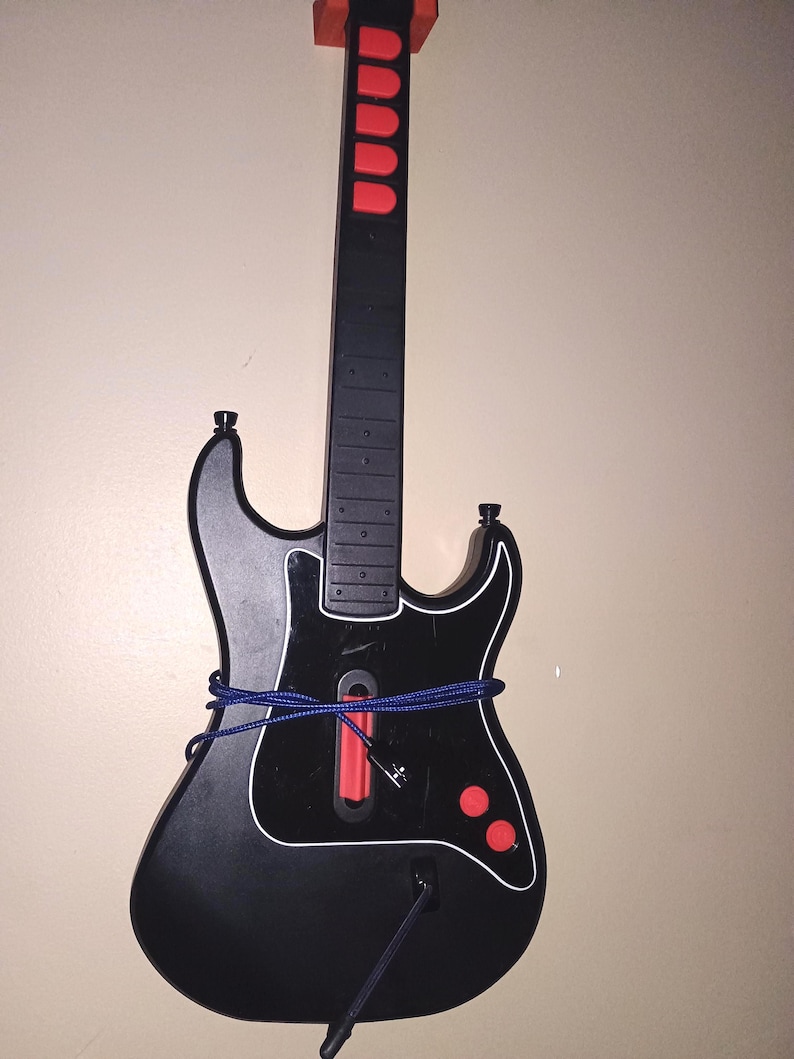 Op de afbeelding: Een zwart-rode gitaarvormige videogamecontroller met witte rand. De controller heeft rode knoppen en een blauw koord eromheen gewikkeld. De bovenkant van de controller is oranje.