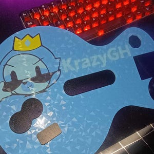 May include: A light blue guitar-shaped cutout with a holographic geometric pattern. It features a cartoon character wearing a yellow crown and black sunglasses, the text KrazyGH, and two black foam-like attachments on the lower section.