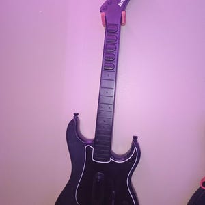 Op de afbeelding: Zwarte Kramer gitaarvormige gamecontroller met witte rand, aan de muur bevestigd. De kop heeft de merknaam. De controller heeft een lange hals met fretmarkeringen en een body met een slagplaat en bedieningselementen.