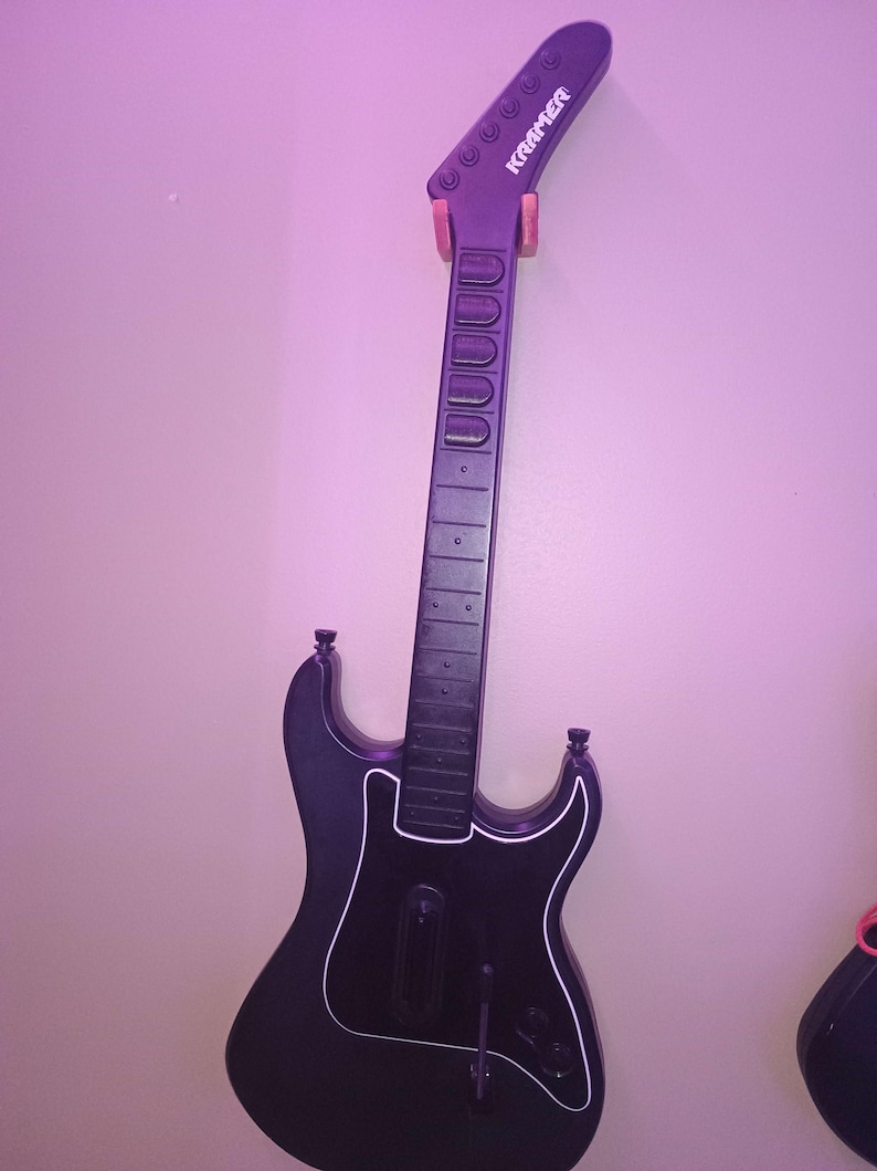 Op de afbeelding: Zwarte Kramer gitaarvormige gamecontroller met witte rand, aan de muur bevestigd. De kop heeft de merknaam. De controller heeft een lange hals met fretmarkeringen en een body met een slagplaat en bedieningselementen.