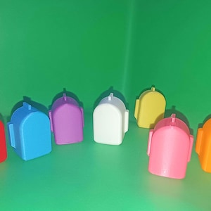 May include: A selection of brightly coloured, 3D-printed mailbox-shaped objects in red, blue, purple, white, yellow, pink, and orange. They are arranged on a green surface.
