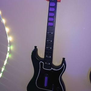 Op de afbeelding: Zwarte speelgoedgitaar met witte rand, paarse knoppen en een rode houder. De gitaar heeft een zwarte hals met zilveren accenten en het woord "Kramer" op de kop. De gitaar is bevestigd aan een lichtpaarse muur.