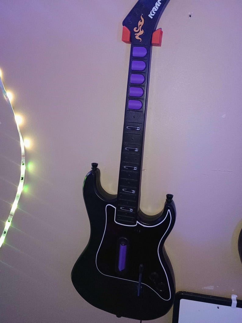 Op de afbeelding: Zwarte speelgoedgitaar met witte rand, paarse knoppen en een rode houder. De gitaar heeft een zwarte hals met zilveren accenten en het woord "Kramer" op de kop. De gitaar is bevestigd aan een lichtpaarse muur.