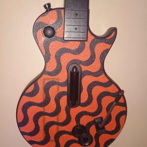 Puede incluir: Un mando de videojuego con forma de guitarra que presenta un patrón ondulado de color naranja y negro en el cuerpo. El mando incluye un mástil negro, una barra de rasgueo central, una palanca de vibrato y botones circulares. La palabra Wavy está en azul.