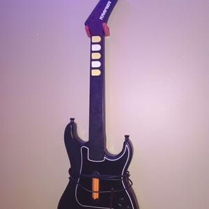 Op de afbeelding: Zwarte gitaarvormige gamecontroller met witte randen en een zwarte hals. De controller heeft een paarse kop met het woord "Kramer" in het wit. Het heeft gele en witte knoppen en een oranje balk.