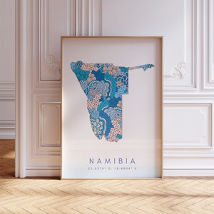 Könnte beinhalten: Ein gerahmter Druck einer Karte von Namibia mit einem blauen und rosa floralen Muster. Die Karte ist von einem goldenen Rahmen umgeben und der Text "NAMIBIA 22.9576° S, 18.4904° E" ist unter der Karte gedruckt.