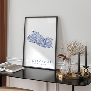 Peut inclure: Une impression encadr&eacute;e en noir avec une carte stylis&eacute;e bleu et blanc du Salvador. La carte est remplie de motifs et de designs complexes. Le texte "EL SALVADOR 13.7942&deg; N, 88.8965&deg; W" est imprim&eacute; sous la carte.