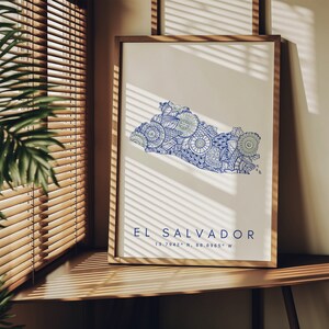 Peut inclure: Une impression encadr&eacute;e d'une carte stylis&eacute;e du Salvador, d&eacute;limit&eacute;e en bleu et remplie de motifs de mandalas complexes. La carte est sur un fond blanc avec le texte "EL SALVADOR 13.7942&deg; N, 88.8965&deg; W" en dessous.