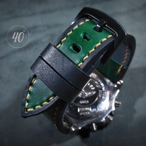 Puede incluir: Una correa de reloj de cuero negro y verde con detalles de costura dorada. La correa tiene un cierre de hebilla y está diseñada para un reloj con una anchura de asas de 40 mm.