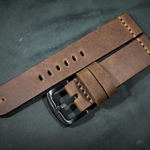 Op de afbeelding: Een bruin leren horlogebandje met een zwarte metalen gesp. Het bandje heeft stiksels langs de randen en gaten om de maat aan te passen.