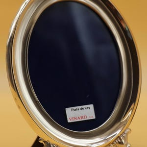 Puede incluir: Marco de fotos ovalado de plata con un respaldo de terciopelo azul. El marco tiene un soporte decorativo con tres patas y una etiqueta que dice "Plata de Ley VINARD, S.A."
