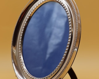 May include: An oval silver picture frame with a polished metallic finish and a beaded inner border. The frame is supported by three ornate, curved feet and features a dark blue insert, standing upright on a small, angled rear support strut.