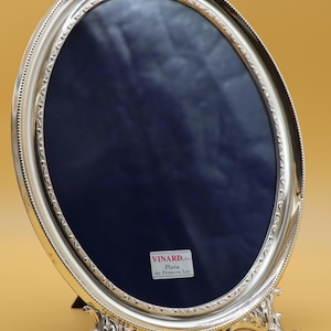 Puede incluir: Un marco de fotos ovalado de plata con un borde exterior de cuentas y un patrón ornamental de hojas a lo largo del borde interior. El marco se apoya sobre dos pies decorativos y contiene un fondo de terciopelo azul oscuro con una etiqueta blanca que dice VINARD, S.A. Plata de Primera Ley.