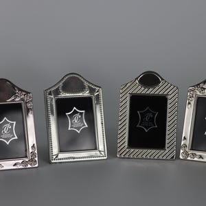 Puede incluir: Cuatro marcos de fotos rectangulares de metal plateado con partes superiores arqueadas. Cada marco presenta un inserto negro con un logotipo blanco que dice JC. Los marcos tienen diseños de borde variados, incluyendo rosas en relieve, bordes con cuentas y rayas diagonales.