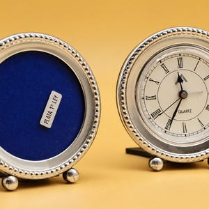 Puede incluir: Dos marcos redondos de tono plateado con bordes de cuentas y pequeños pies esféricos. Un marco contiene una esfera de reloj con números romanos negros y la palabra QUARTZ. El otro tiene un fondo de terciopelo azul con la etiqueta PLATA 1ª LEY.