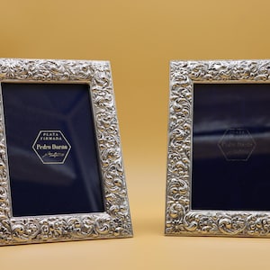 Puede incluir: Dos marcos de fotos plateados con detalles florales ornamentados. Cada marco tiene un interior azul oscuro y una etiqueta que dice "Plata Firmada Pedro Duran". Los marcos se muestran sobre una superficie marrón claro.