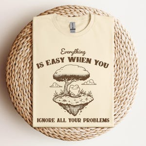 May include: Cream-colored t-shirt with a graphic featuring a tree, a frog, and the text "Everything is easy when you ignore all your problems." The design is in shades of brown and the shirt has a crew neck.
