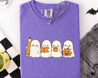 May include: A purple Comfort Colors t-shirt featuring a graphic of four cartoon ghosts, each holding a pencil, book, mug, and pumpkin. A silver chain necklace is draped across the neckline. The shirt is folded on a white surface.