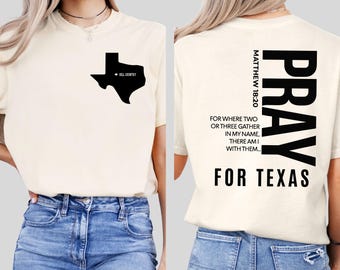 May include: Cream-colored t-shirt with a black Texas outline and "Hill Country" text. The back features the word "PRAY" vertically, Bible verse Matthew 18:20, and "FOR TEXAS" in bold black letters. The shirt is paired with blue jeans.