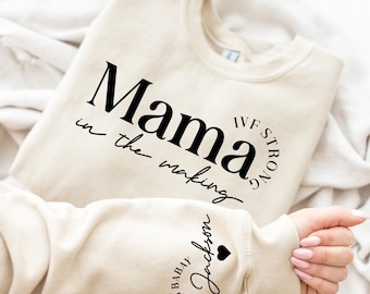 以下が含まれることがあります： ベージュのトレーナーに、「Mama in the making IVF Strong」という黒い文字が書かれています。袖には、「Making Baby Jackson」という黒い文字と小さなハートが書かれています。