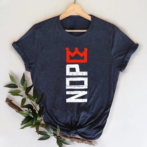 May include: A navy blue t-shirt with a red crown graphic above the white letters "NDP" in a vertical arrangement. The t-shirt is hanging on a wooden hanger.