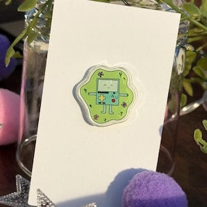 May include: A green and white sticker featuring a cartoon character resembling a retro handheld game console. The sticker has a green outline with floral and leaf accents. The character has a smiling face and a cross-shaped control pad.