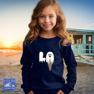Op de afbeelding: Marineblauw sweatshirt met lange mouwen met een witte afbeelding van de letters "LA", een smeltend spook en een ijshoorntje. Het sweatshirt wordt gedragen door een jong persoon op een strand met een strandwachtpost op de achtergrond.