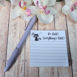 May include: A light purple ballpoint pen next to a square white notepad with horizontal lines. The notepad features a black graphic of a wide-eyed cat and the printed text, It's fine! Everything's fine! The pen has a textured grip area.
