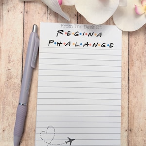May include: A lined notepad with the text "From The Desk Of: REGINA PHALANGE" in a colorful font, with a heart and airplane graphic. A lavender pen rests beside the notepad. White and pink orchid flowers are in the background.