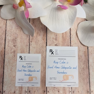 May include: Two prescription-style cards with the text "Keep Calm + Send Home Gabapentin and Trazodone" in blue script. The cards feature a pill bottle illustration and are set against a wooden background, with white and pink orchid flowers.