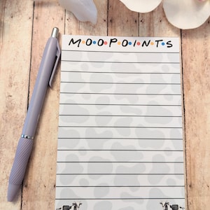 May include: A lined notepad with the text "MOO POINTS" in black, with each letter in a different color. The notepad has a cow print design and two cow illustrations at the bottom. A lavender pen rests on the side of the notepad.