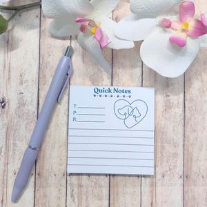 May include: A light purple pen rests beside a white notepad with the words "Quick Notes" and a heart-shaped dog and cat design. The notepad has lined spaces for notes. White orchid flowers with pink accents are in the background.