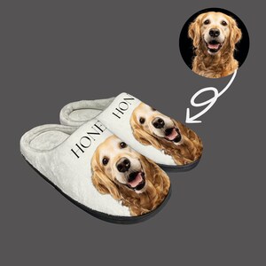 May include: A pair of white fleece slippers with a black border. Each slipper features a golden retriever dog face with the word "HOME" printed on the side.