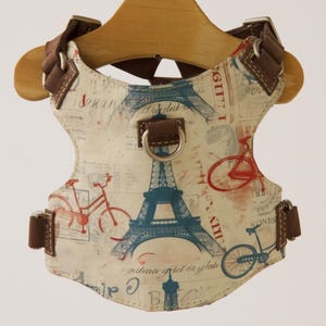 May include: Dog harness with a cream background, decorated with blue Eiffel Towers and red bicycles. The harness has brown leather straps and a metal D-ring. The harness is designed with a Parisian theme.