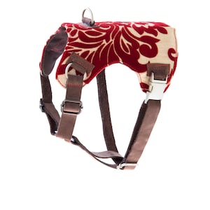 May include: A dog harness with a red and beige floral design. It features brown leather straps, metal fixings, and a D-ring for leash attachment. The harness includes a buckle closure for a secure fit.