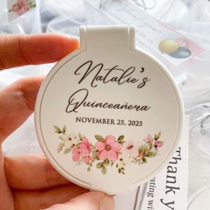 Peut inclure: Un miroir de poche blanc avec le texte "Natalie's Quinceañera" et la date du 25 novembre 2025. Le miroir est décoré d'un motif floral rose et blanc. Le miroir est tenu dans une main.