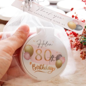 May include: A white, round party favor with the text "Helen 80th Birthday" in gold script, with pink and gold balloon graphics. The favor is in a sheer white bag, with a tag that says "Thank you for celebrating with us."